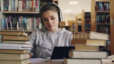 戴着白色耳机的积极微笑的年轻女子坐在<strong>大学图书馆</strong>的桌子旁，手里拿着数码平板电脑和